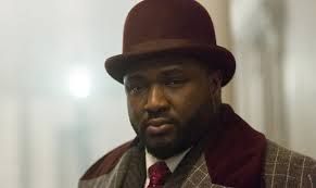 Nonso Anozie wears a red bowler hat and smartly tailored grey check suit, with a tie that matches his hat as Renfield