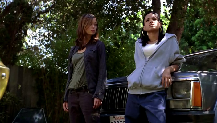 Cameron and Chola leaning on a car, Cameron stares directly at her while Chola affects nonchalence