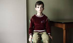 Victor is a little pale boy in a red jumper with a cowlick hair style sitting on a chair looking straight at the camera with