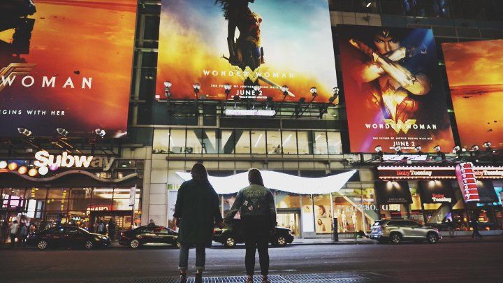 Image shows three huge Wonder Woman posters hanging over a cinema. Two women stand in front of the cinema holding hands
