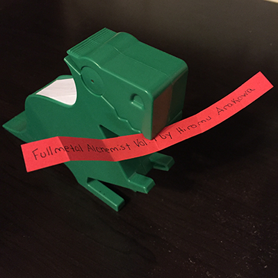 Green dinosaur holding orange slip of paper reading Fullmetal Alchemist by Hiromu Arakawa