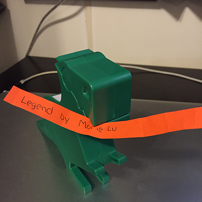Green dinosaur holding orange slip of paper reading Marie Lu by Marie Lu