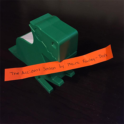Green dinosaur holding orange slip of paper reading The Accident Season by Moïra Fowley-Doyle